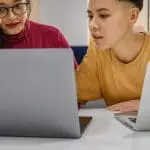 Two people wearing bright clothes looking at a laptop and speaking