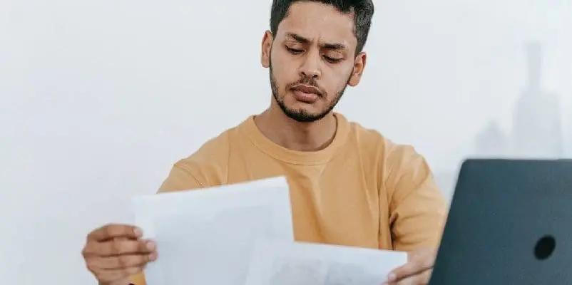 Man looking through study notes, sitting in front of laptop