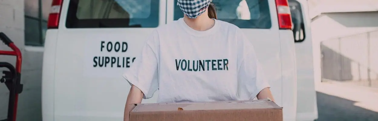 person in a mask wearing a volunteer t shirt and holding a cardboard box labelled food