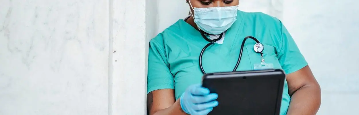 healthcare worker in scrubs and mask holding a tablet computer and looking at it