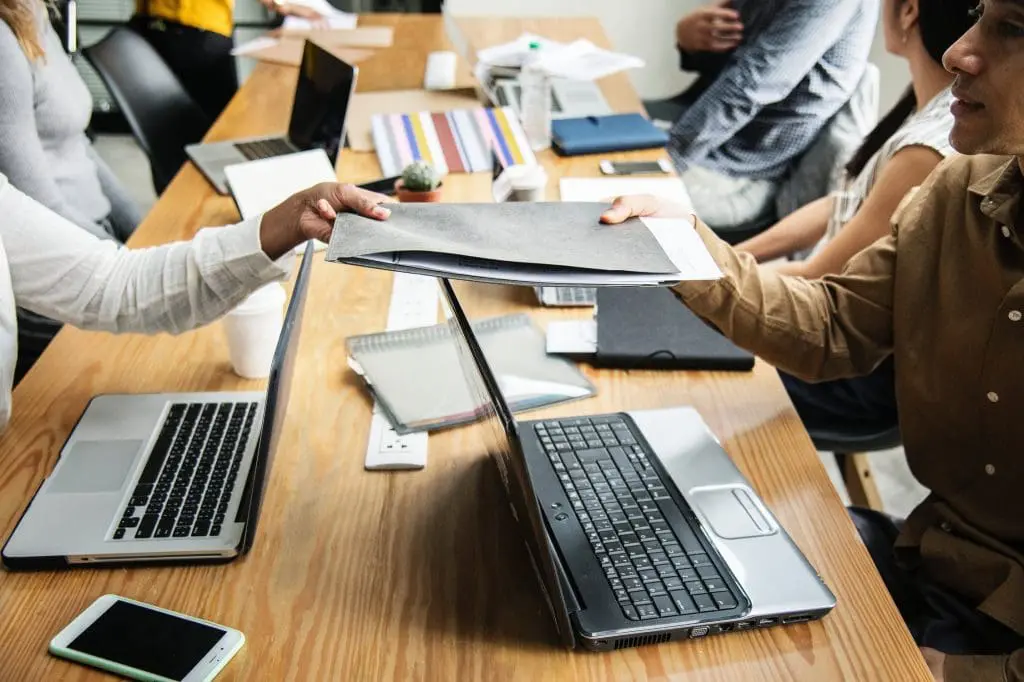 office workers sat at a meeting table filled with laptops with a focus on two colleagues exchanging a physical file.