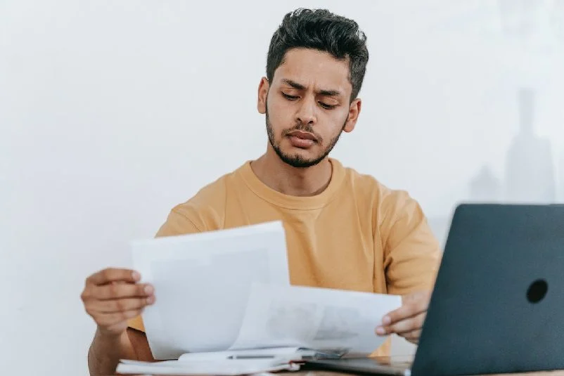 Man looking through study notes, sitting in front of laptop