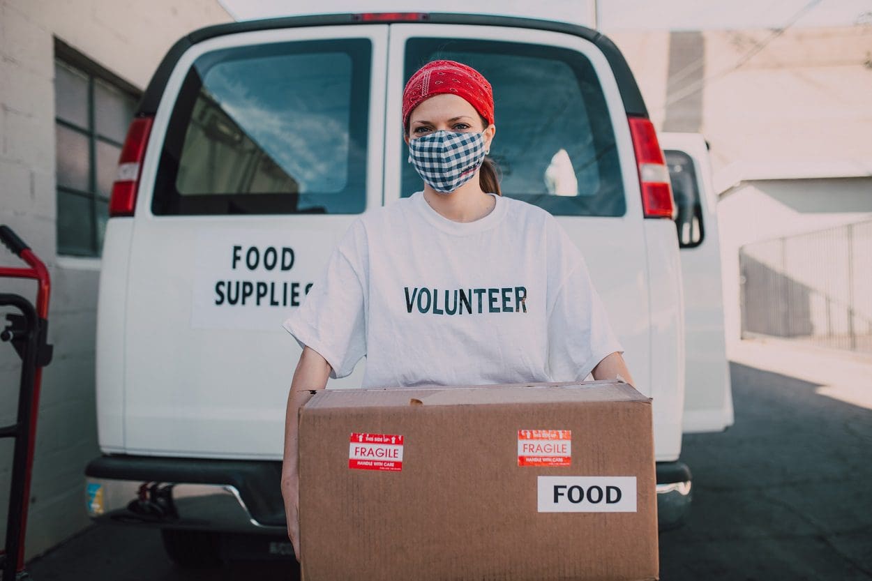 person in a mask wearing a volunteer t shirt and holding a cardboard box labelled food